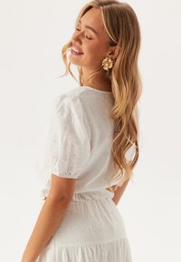 Blusa bianca con maniche a palloncino e ricami floreali. Il tessuto è leggero con una finitura texture. Indossata con una gonna abbinata.