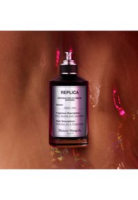 Flacon de parfum en verre noir avec un vaporisateur argenté. L'étiquette inclut "REPLICA" et les détails de la fragrance, reposant sur une surface brillante.