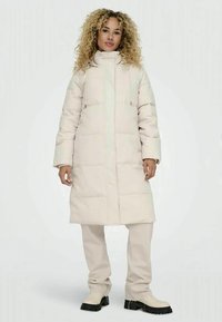 Mulher com cabelo encaracolado, vestindo um longo casaco acolchoado bege, calças a condizer e botas claras com solas pretas, de pé contra um fundo plain.