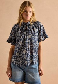 Námorníková blúzka s kvetinovým vzorom, krátkymi volánovými rukávmi, výrezovým výstrihom a textúrovanou látkou. Spárovaná s modrými džínovými šortkami.