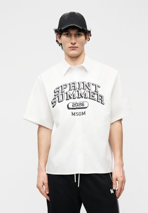 Jaunas vyras dėvi juodą kepuraitę, baltą trumpomis rankovėmis marškinėlį su užrašu „SPRINT SUMMER 2026 MSGM“ ir juodas kelnes su baltais raišteliais.