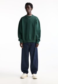 Green oversized sweatshirt with ribbed cuffs, paired with loose dark denim jeans and white sneakers. Silver chain necklace detail visible.