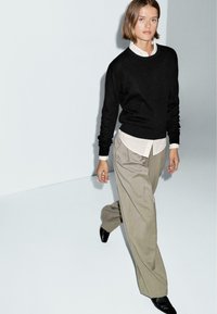 Maglione nero sopra una camicia bianca con colletto, abbinato a pantaloni beige chiaro su misura. L'outfit presenta una vestibilità comoda e texture morbide.