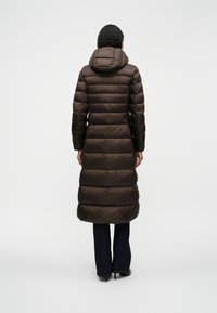 Lang brun dunjakke med hætte, prydet med et quiltet design, glat tekstur og en tætsiddende silhuet. Båret over mørke jeans.