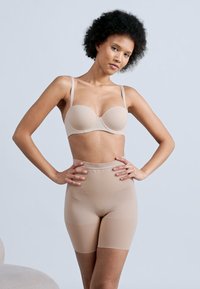 Γυναίκα με σγουρά μαλλιά, ποζάρει με τα χέρια στη μέση φορώντας μπεζ στηθόδεσμο χωρίς λουράκια και ψηλοτάτη αόρατη σμιλευτική βερμούδα, πάνω σε γκρίζο φόντο.