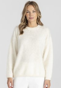 Kobieta o falujących blond włosach, ubrana w miękki, puszysty sweter w jasnym odcieniu beżu i białe spodnie, stojąca na tle jednolitego, jasnego tła.
