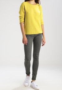 Gelbes, strukturiertes Sweatshirt mit Rundhalsausschnitt, kombiniert mit grauen, eng anliegenden Jeans. Weiße Sneakers runden das Outfit ab.