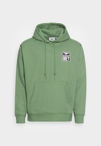 Grön sweatshirt med huva och ficka fram, samt en liten svart-vit "OBEY"-logotyp på vänster bröst, med justerbara snören på huvan.