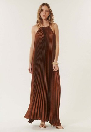 Femme portant une robe maxi plissée marron sans manches et des sandales à talons hauts dorées, debout avec une expression neutre sur un fond uni.