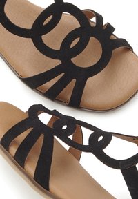 Black suede sandals featuring a cut-out design with circular accents and straps. The footbed is a light tan color, providing contrast.