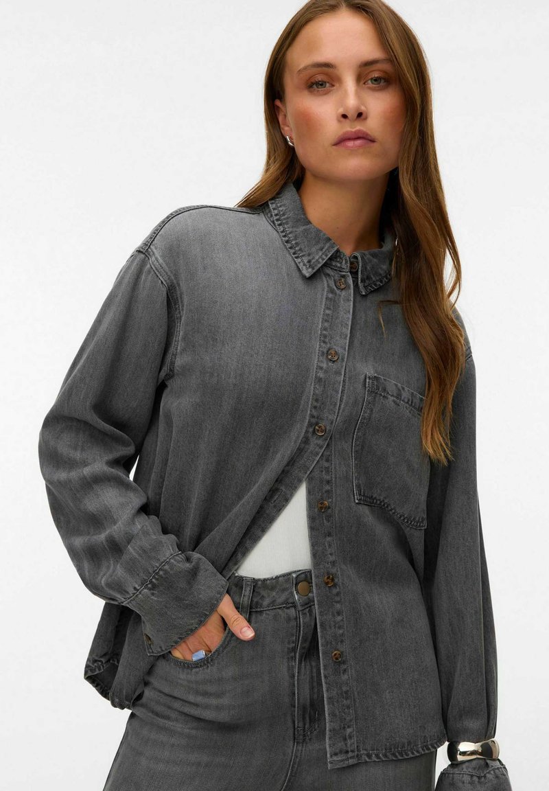 Jeune femme aux longs cheveux bruns porte une chemise en jean gris foncé ample et un jean assorti, une main dans sa poche.
