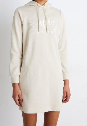 Femme portant une robe sweat à capuche à manches longues crème avec le logo "GANT American Sportswear 1949" sur la poitrine.