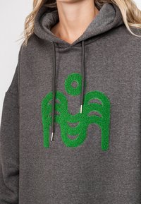 Harmaa huppari, jossa on etupuolella teksturoitu vihreä logo. Hupparissa on naruhuppu ja se on valmistettu pehmeästä, lämpimästä kankaasta.