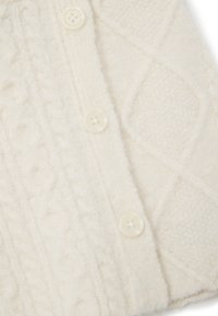 Gilet en tricot couleur crème avec des motifs en relief en câble, doté de trois boutons blancs et d'un tissu doux et duveteux.