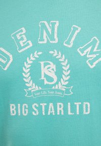 Tirkīza kokvilnas t-krekls ar baltu novecojošu uzrakstu, apļveida dizainu ar lauru lapām un treknu tekstu: "DENIM BIG STAR LTD."