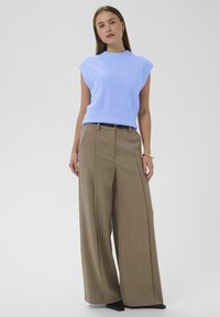 Nainen pukeutuneena vaaleansiniseen ilman hihoja neulepaitaan, leveisiin beigeihin housuihin mustalla vyöllä, kultaketjuun ja mustiin kärjellisiin kenkiin, seisomassa valkoista taustaa vasten.