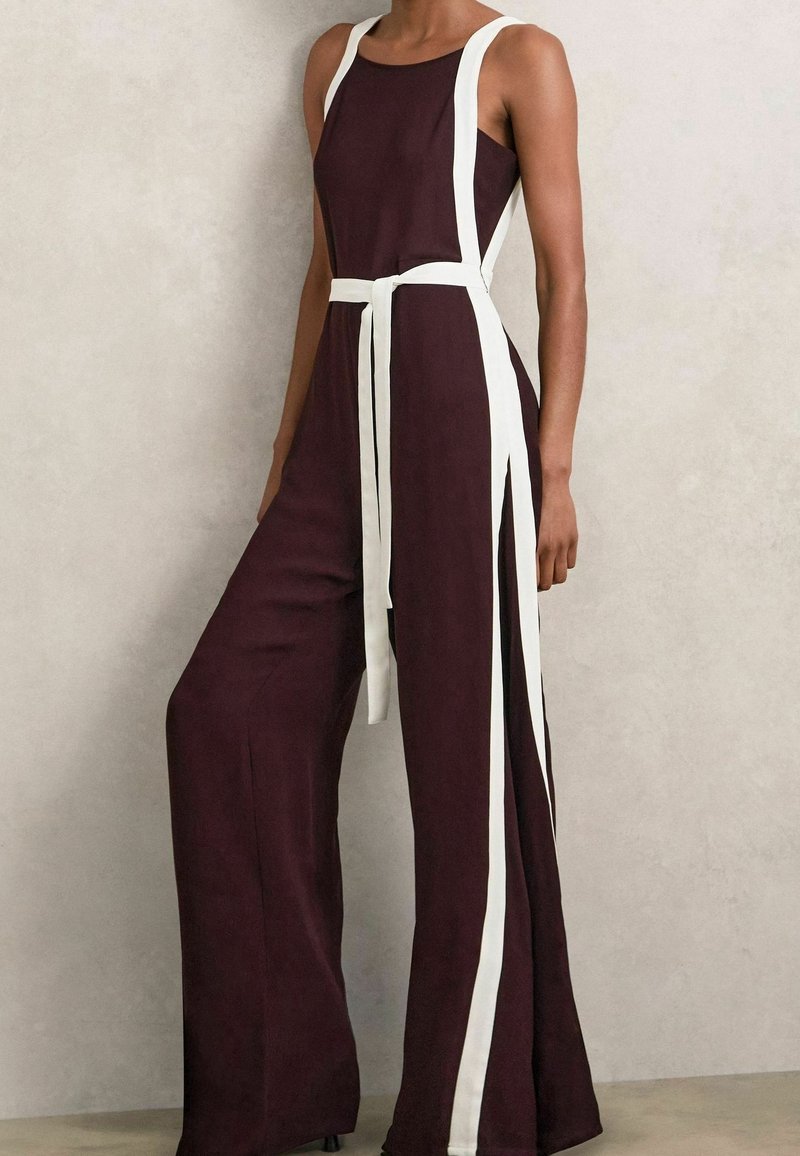 Combinaison sans manches violet foncé avec jambes larges, rayures verticales blanches sur les côtés et ceinture blanche nouée à la taille.