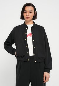 Μαύρο μπουφάν σε στιλ μπότερ από απαλό ύφασμα, με στρογγυλή λαιμόκοψη, κλείσιμο με κουμπιά τύπου σκρατς και δύο τσέπες μπροστά. Σχεδιασμένο για άνετη εφαρμογή.