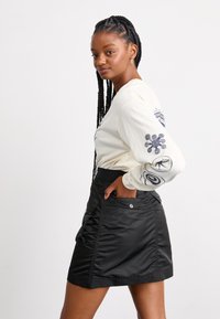 Black, knee-length skirt with a textured finish and side pockets. Paired with a light cream long-sleeve top featuring graphic designs on sleeves.