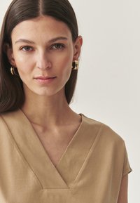 Beige, V-nyakú felső rövid ujjakkal, sima anyagból készült, tiszta vonalú és strukturált nyakkivágással. Arany karika fülbevalók.