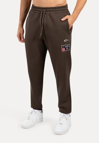 Pantalones de chándal marrones con una cinturilla elástica, bolsillos laterales y un detalle de parche. Combinados con zapatillas blancas. Textura de tela suave.