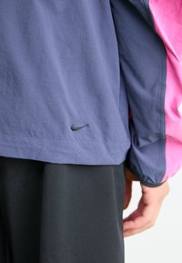 Nike jakke i blå stof med pink detaljer, med et diskret logo på den nederste kant. Glat tekstur med en afslappet pasform.