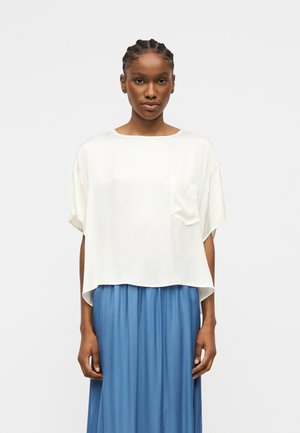 Femeie cu păr împletit poartă un top alb larg cu mâneci scurte și buzunar, și o fustă plisată albastră cu talie înaltă, stând în fața unui fundal simplu.