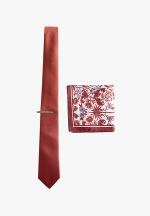 Red textured necktie with silver tie clip beside folded white pocket square with red and blue floral pattern.