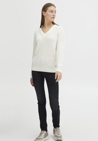 Strikket elfenbensfarget genser med V-hals, lange ermer og ribbestrikkede mansjetter. Kombinert med svarte skinny jeans og lysegrå joggesko.
