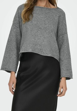 Femme portant un pull en maille gris ample à larges manches et une jupe en satin noire, vue du cou aux hanches sur un fond neutre.