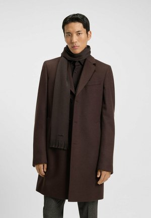 Cappotto di lana marrone con un design su misura, abbinato a una sciarpa marrone coordinata. La sciarpa presenta dettagli frangiati ed è indossata sopra una maglietta nera.