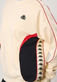 Béžová mikina Kappa s červenými lemami, čiernymi bočnými panelmi a opakujúcim sa čiernym logom Kappa na krémovej stužke na rukáve.