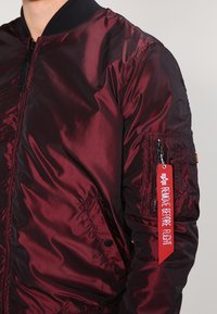 Burgundowa kurtka typu bomber z błyszczącego, lekkiego materiału, z prążkowanym kołnierzem, bocznymi kieszeniami oraz zapinaną na zamek kieszenią na rękawie z czerwonym oznaczeniem.