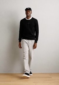 Felpa nera con logo, jeans grigi chiari e sneakers nere con suola bianca, abbinate a un cappellino da baseball nero. Sfondo in legno.
