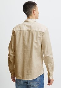 Camisa de manga larga beige hecha de tejido ligero, con cuello extendido y puños abotonados, combinada con jeans de mezclilla azules.