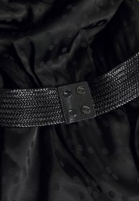 Černý tkaný pásek s obdélníkovou sponou připomínající kůži, která má dvě lesklé kulaté cvoky. Pozadí tvoří černá látka s puntíkovaným vzorem.