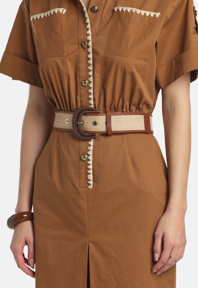 Bruine jurk met korte mouwen, decoratieve crèmekleurige bies, knopen aan de voorkant, brede beige en bruine riem, en een armband om de linkerpols.