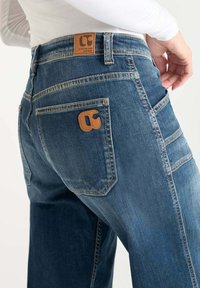 Jeans de mezclilla azul con cintura alta, parche de logo en cuero color tan en la parte trasera, y tres bolsillos traseros con detalles de costura en contraste.