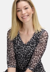 Lächelnde Frau mit blonden Haaren, die ein durchsichtiges, schwarz-graues Leopardenmuster-Top trägt, vor einem schlichten hellen Hintergrund.