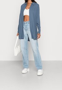 Chemise à manches longues, boutonnée en tissu bleu, associée à un crop top blanc et un jean taille haute bleu clair. Sac à main blanc et chaussures à plateforme.