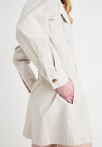 Beige bomulls/kjole med lange ermer, kneppede mansjetter og sidelommer. Har en tettsittende midje og rett snitt. Minimalistisk design.