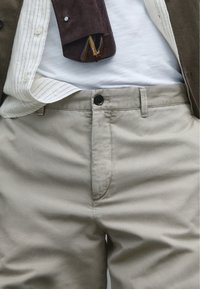 Kaki-Hosen mit Knopfverschluss, ausgestattet mit Gürtelschlaufen, frontalen Taschen und einer glatten Textur. Eine braune Wildledertasche ist teilweise sichtbar.