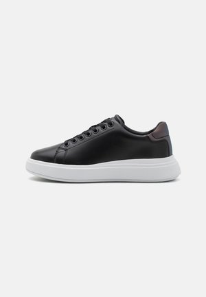 Sneaker basse nere con lacci neri e colletto imbottito, caratterizzata da una spessa suola in gomma bianca su uno sfondo chiaro e semplice.