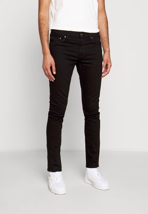 Hombre vestido con una camiseta blanca y jeans negros, de pie frente a un fondo neutro, acompañado de zapatillas blancas.