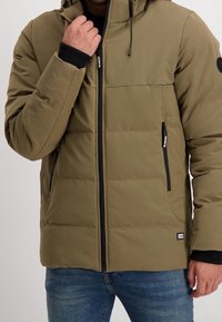 Olivgrön puffjacka med huva, blixtlås framtill, svarta detaljer och två sidofickor. Har quiltat foder för isolering.