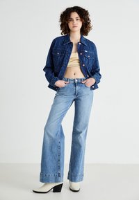 Chaqueta de denim en azul oscuro con patrón texturizado, dos bolsillos frontales; combinada con pantalones de mezclilla de pierna ancha en azul claro, botas de tacón blancas y top corto dorado.