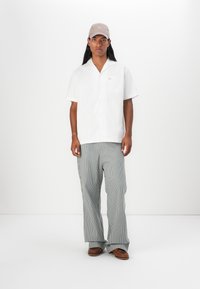 Camicia bianca a maniche corte con taschino, abbinata a pantaloni a gamba larga grigio chiaro a righe e scarpe marroni, completata da un cappello beige.