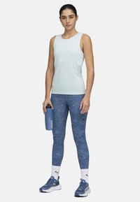 Világoskék ujjatlan top, kék mintás leggings, vízpalackot tart, fehér zoknik logóval, kék sportcipők.