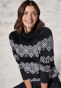 Zwarte fuzzy trui met een col, voorzien van een wit geometrisch patroon. Lange mouwen en een zachte textuur, gecombineerd met donkere jeans.