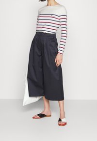 Gestreiftes Langarmoberteil in Weiß, Marineblau und Rot; kombiniert mit weiten schwarzen Culottes und schwarz-weißen RutschSandalen, glatte Texturen sind sichtbar.